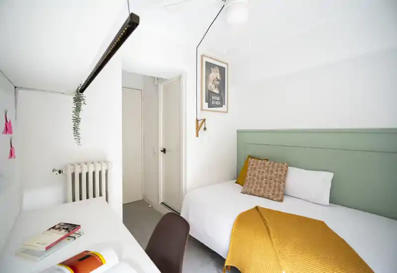 Camera da letto compatta e luminosa con letto singolo contro una testiera verde pastello, coperta ocra e cuscino decorato. Piccola scrivania e sedia, pareti bianche pulite e illuminazione moderna minimal.