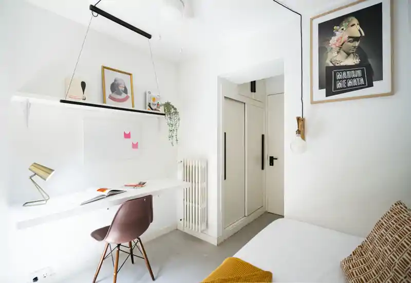 Camera da letto luminosa e minimalista con scrivania bianca, sedia moderna e letto parzialmente visibile con coperta e cuscino accoglienti; quadri e mensole aggiungono carattere contemporaneo.