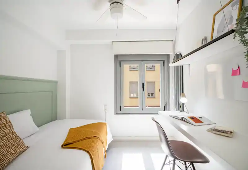 Camera da letto luminosa e minimalista con letto confortevole, tessili accoglienti e una scrivania integrata vicino alla finestra — ideale per un soggiorno compatto e funzionale.