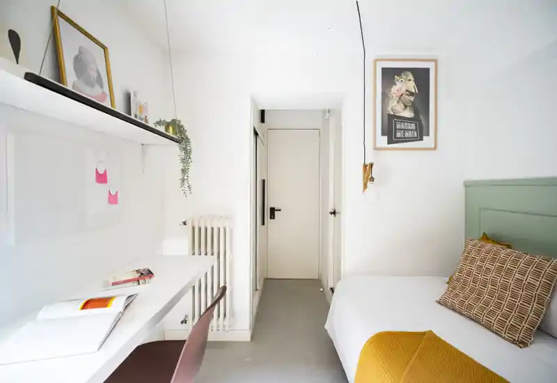 Camera da letto luminosa e compatta con un letto singolo ben rifatto contro una testiera verde pastello, cuscino stampato e coperta senape. Una scrivania bianca con mensole e una sedia corre lungo la parete sinistra, creando una zona lavoro funzionale.