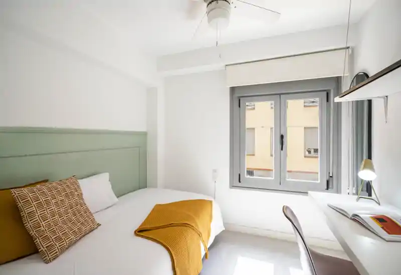 Camera da letto luminosa e moderna con letto ben rifatto, testata verde salvia e accenti color senape. Una piccola scrivania vicino alla finestra offre uno spazio di lavoro funzionale e molta luce naturale.