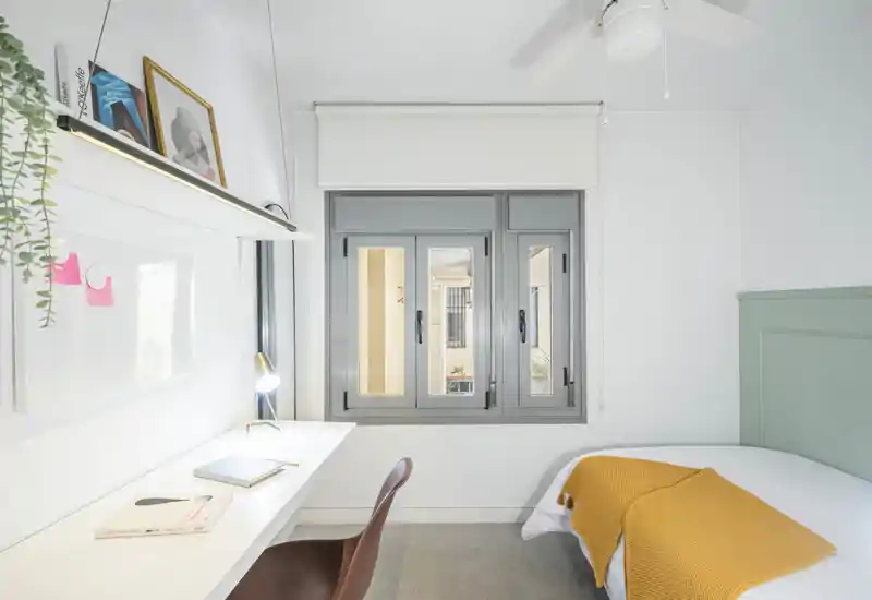 Bright, minimal bedroom with a single bed dressed with a mustard throw, a built-in desk and large window providing natural light — ideal for a student or single occupant.