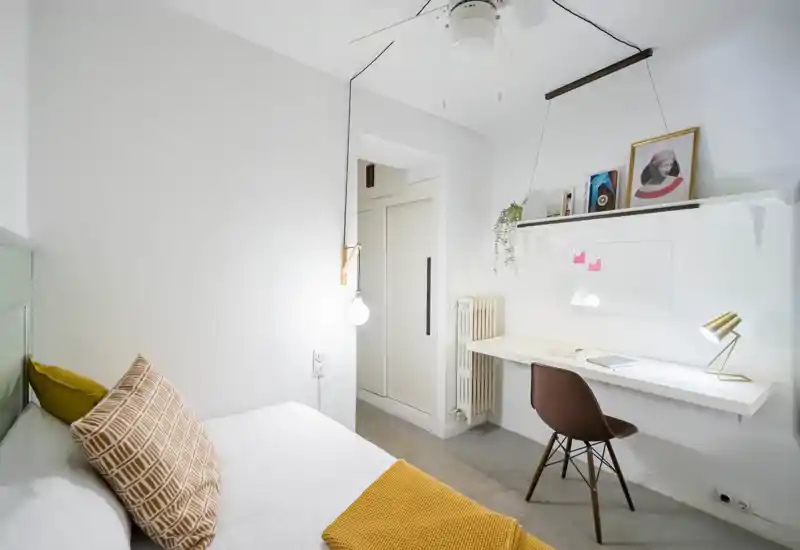 Dormitorio pequeño y luminoso con cama individual, cojines decorativos y una manta mostaza. Escritorio mural minimalista y silla que crean un espacio de trabajo ordenado; paredes blancas y luz natural aportan frescura.