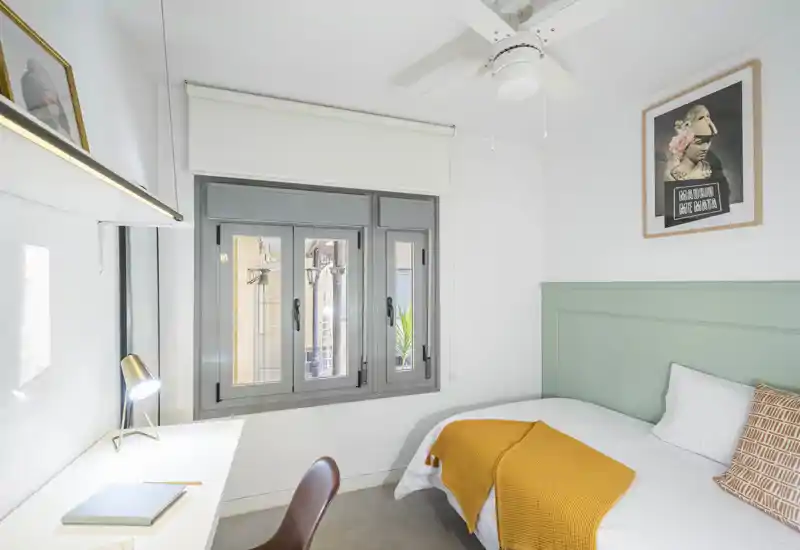 Bright, neatly styled bedroom with a comfortable single bed, soft green headboard and a warm mustard throw. The space includes a compact desk by the window, ceiling fan and modern decorative artwork—ideal for a cozy, well-lit rental.