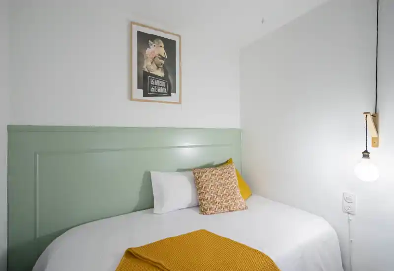Bright, neatly styled single bedroom with a soft green headboard, white bedding and warm mustard accents. The room feels fresh and welcoming with clean lines and a minimalist pendant light.