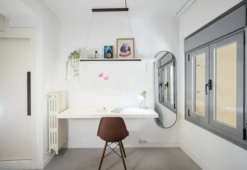 Espacio de trabajo en casa luminoso y minimalista con escritorio de pared, silla moderna, lámpara de escritorio y estante decorativo; amplias ventanas aportan luz natural, ideal para concentrarse o estudiar.