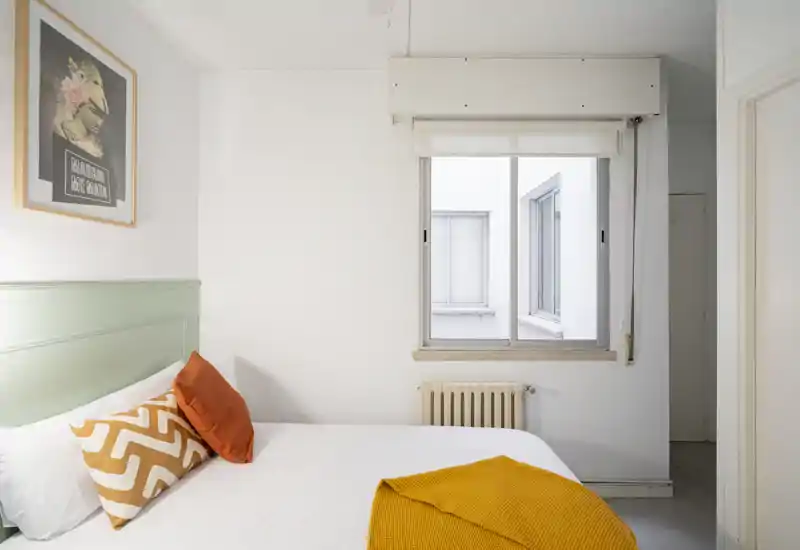 Chambre lumineuse et minimaliste avec un lit bien fait, des coussins décoratifs et un plaid moutarde, et une fenêtre apportant de la lumière naturelle.