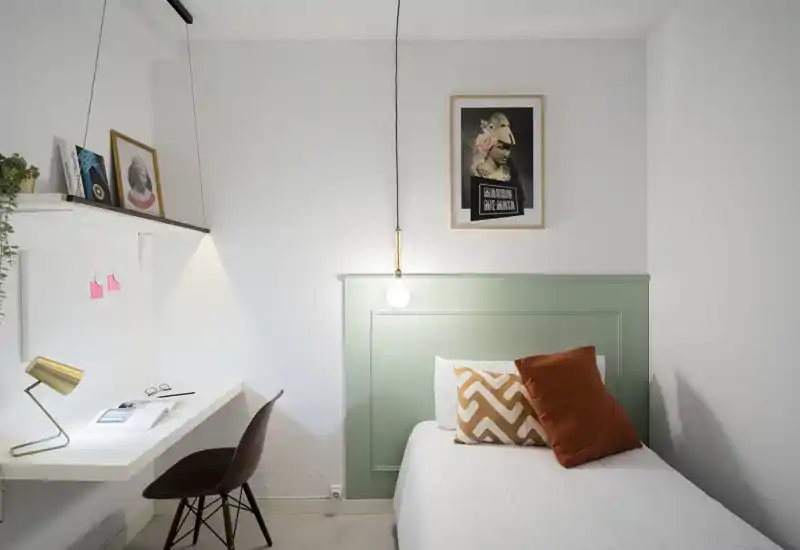 Chambre simple compacte et stylée avec une tête de lit vert menthe, literie blanche et coussins chauds ; coin bureau avec lampe et étagères, parfaite pour une personne seule.