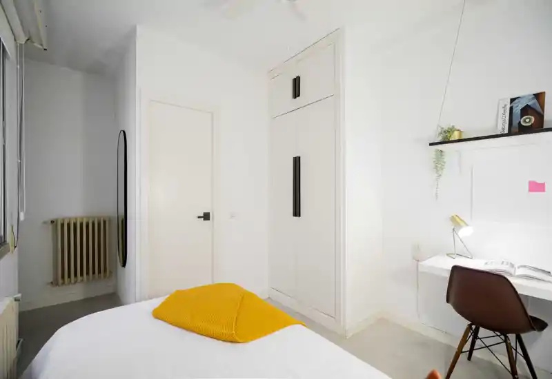 Chambre lumineuse et minimaliste avec lit bien fait et plaid jaune, armoire encastrée et petit bureau avec lampe — parfaite pour courts séjours ou voyageurs seuls.