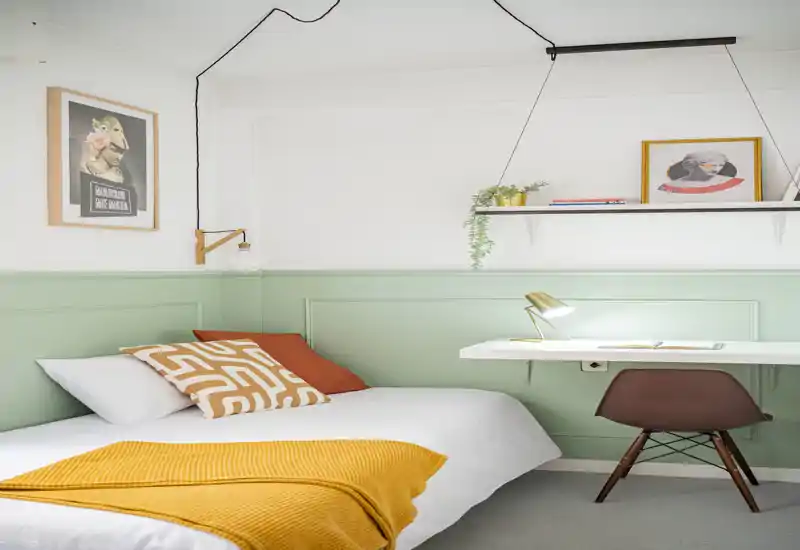 Bright, stylish bedroom with a white bed, patterned cushions and a mustard throw. A compact workspace with a floating desk and lamp sits next to the bed, with decorative shelves and artwork above.