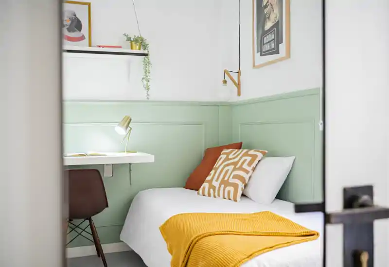 Bright, stylish single bedroom with a neatly made bed, warm mustard throw and decorative cushions against a mint-green paneled wall; compact workspace with a wall-mounted desk and lamp.