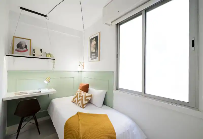 Bright, compact bedroom with a single bed dressed in white linen, warm ochre throw and patterned cushion; a small desk and lamp create a practical workspace by the window.
