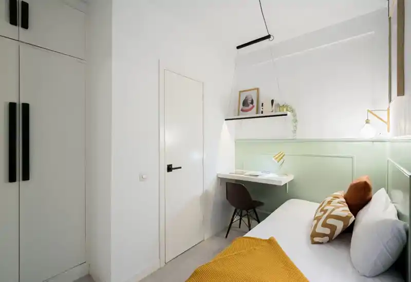 Compact, well-styled bedroom with a comfortable single bed, decorative pillows and a warm throw; a small wall-mounted desk and shelf make it ideal for working or studying.