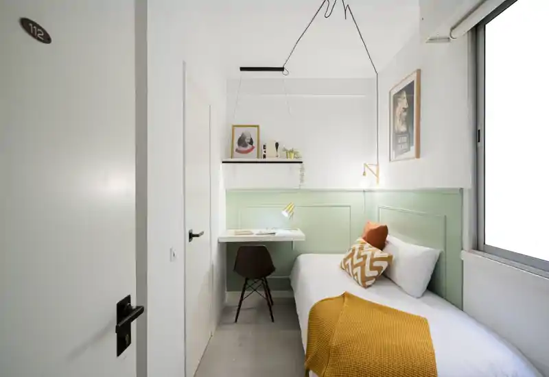 Bright, compact bedroom with a single bed dressed in white linen, warm mustard throw and decorative cushions, plus a small built-in desk by the headboard and large window for natural light.