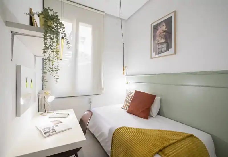 Chambre simple et lumineuse avec lit une place, coussins décoratifs et plaid moutarde chaleureux ; un bureau blanc sous la fenêtre offre un espace de travail soigné et des plantes suspendues apportent une touche fraîche.