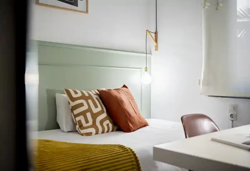 Chambre lumineuse et élégante avec tête de lit vert pâle, coussins décoratifs et suspension lumineuse chaleureuse — un espace de couchage accueillant et soigné pour les annonces.