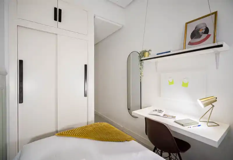 Chambre à coucher compacte et confortable avec lit simple bien fait, placard intégré et petit bureau mural avec lampe — lumineuse, moderne et chaleureuse.