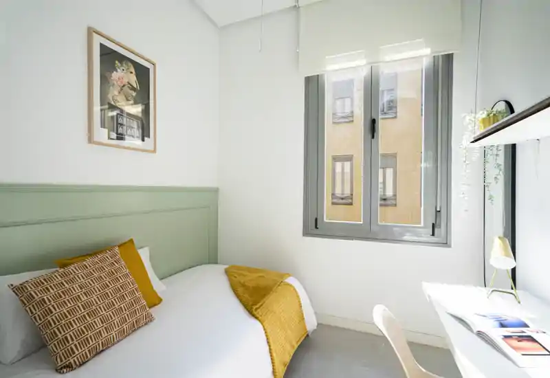 Chambre simple lumineuse et soignée avec une tête de lit verte, accents moutarde et une fenêtre ensoleillée apportant la lumière naturelle.