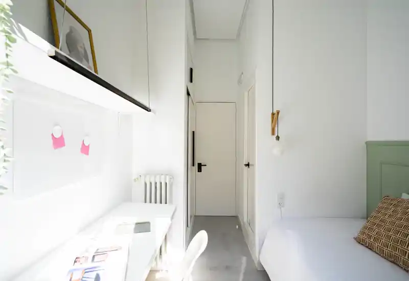 Chambre lumineuse et minimaliste avec un lit blanc, un coussin à motif et un bureau près de la fenêtre ; lignes nettes et lumière naturelle abondante pour une atmosphère aérienne.