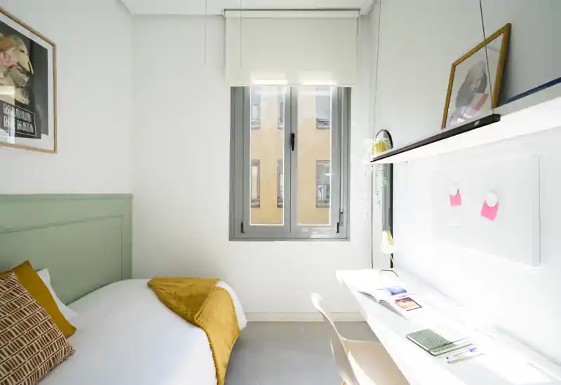 Chambre lumineuse et minimaliste avec un lit simple en linge blanc et un plaid moutarde ; un bureau compact sous une étagère près de la fenêtre, parfait pour étudier ou télétravailler.