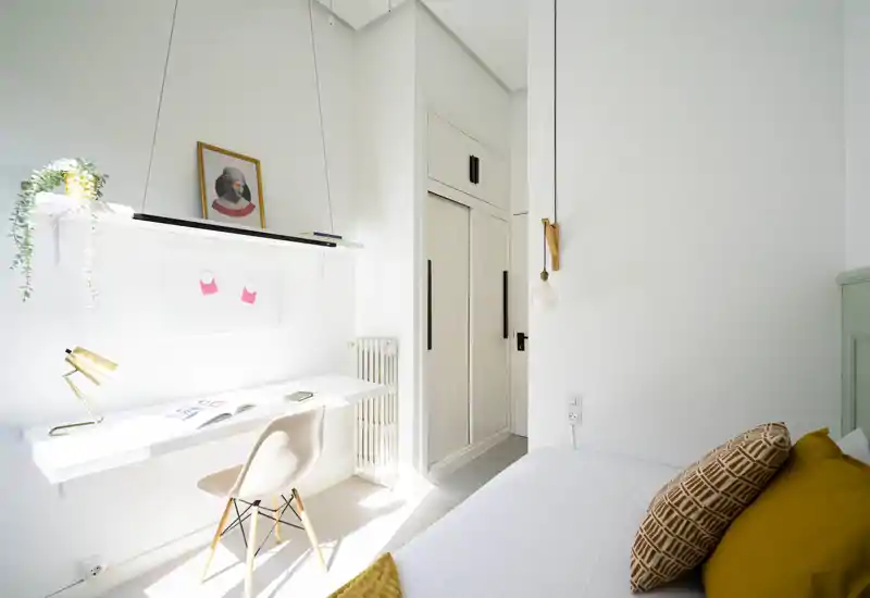 Chambre claire et minimaliste avec un bureau blanc près de la fenêtre, coussins chaleureux et une suspension sobre — idéale pour une location compacte et stylée.