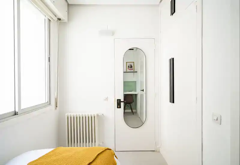 Rincón de dormitorio minimalista y luminoso con paleta blanca y una manta amarilla en la cama; un espejo en la puerta refleja una zona de trabajo contigua.