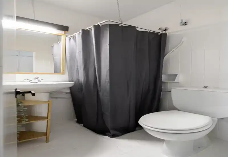 Baño limpio y luminoso con ducha oculta tras una cortina oscura, azulejos blancos, lavabo con espejo y aseo — bien iluminado y de estilo sencillo, ideal para anuncios.