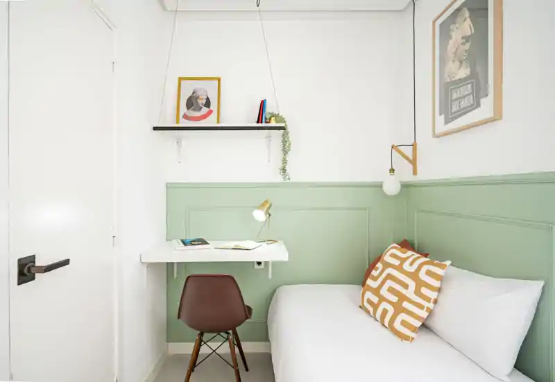 Compact, stylish bedroom with a single bed against a mint-green paneled wall, a white floating desk with lamp, and decorative cushion and artwork for a cozy, modern feel.