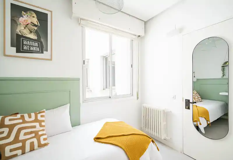 Bright, stylish single bedroom with a green paneled headboard, patterned cushion and mustard throw. Large window and wall mirror give a sense of space and light.