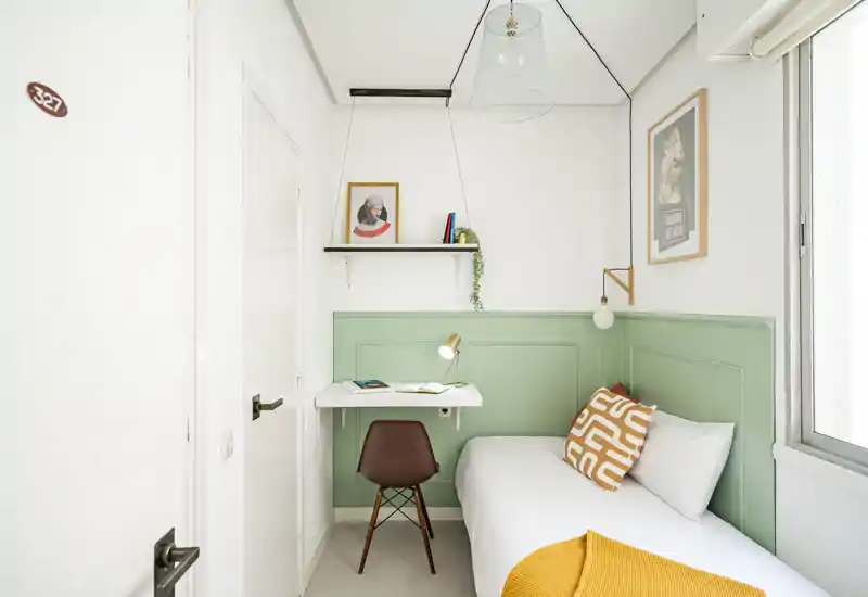 Compact, bright single bedroom with a neatly made bed, bedside lamp and a small wall-mounted desk — ideal for a student or short-stay guest.