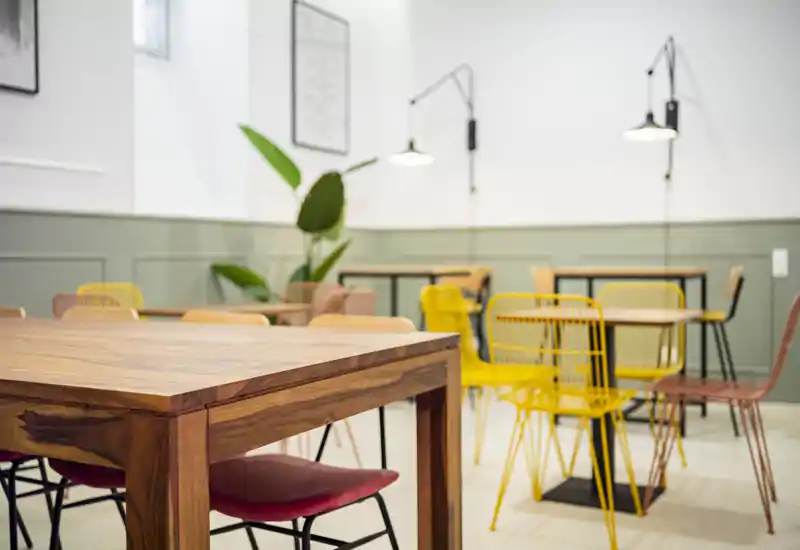 Área de comedor moderna y luminosa con mesa de madera maciza, sillas de colores variadas y lámparas de pared minimalistas — un lugar acogedor para comer o trabajar.