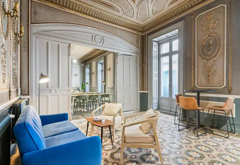 Sala de estar luminosa y elegante con sofá azul, butacas y ventanas altas enmarcadas por molduras ornamentadas y suelo de baldosas decorativas — atractiva y acogedora para socializar.