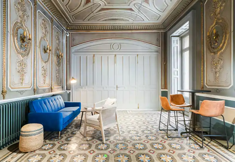Sala elegante con molduras clásicas, sofá azul de terciopelo, asientos variados y suelo de baldosas decoradas; espacio luminoso y con estilo, ideal para relajarse o recibir invitados.
