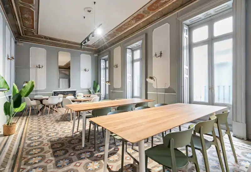 Elegante sala común para comer o reunirse con largas mesas de madera, sillas modernas y altas ventanas que inundan el espacio de luz natural; techo ornamentado y suelo de baldosas con mucho carácter.