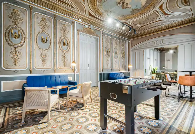 Amplia y elegante sala con molduras decorativas, suelo de baldosas con patrones, asientos cómodos y una mesa de futbolín — un salón común luminoso y con estilo.