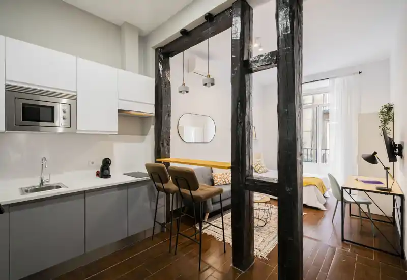Acogedora zona de estar de concepto abierto con cocina compacta, taburetes, sofá y un rincón para dormir muy luminoso, perfecta para un estudio moderno.