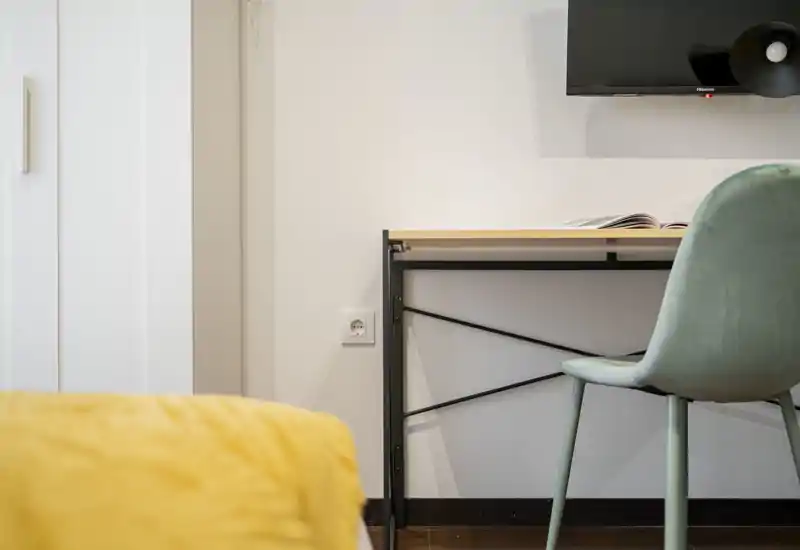 Bright, minimal bedroom corner with a visible yellow duvet in the foreground, a small desk and chair against a pale wall, and a wall-mounted TV. Clean, modern styling with soft natural light.