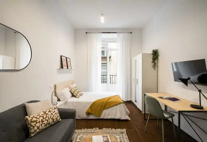 Well-styled bedroom with a double bed centered by a bright window, warm textiles, a small sofa and a desk—ideal for a comfortable stay with natural light and tasteful decor.