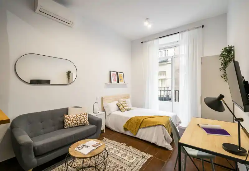 Bright, stylish bedroom with a comfortable single bed, cozy textiles and a small seating area. Natural light from the balcony window enhances the warm wooden floor and modern furnishings.
