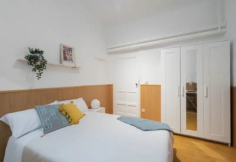 Bright, neatly styled bedroom with a double bed, decorative cushions, warm wood paneling and a white wardrobe with mirrored door—ideal for rental listings highlighting comfort and storage.