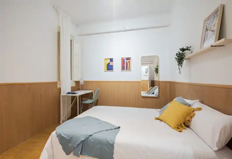 Bright, stylish bedroom with a double bed dressed in white linens, colorful cushions and a soft throw. The room features a small desk by the window, wall art and a full-length mirror that enhances the sense of space.