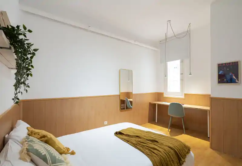 Bright, modern bedroom with warm wood paneling, a neatly made bed with cushions and throw, a desk by the window and minimalist decor — ideal for comfortable stays.