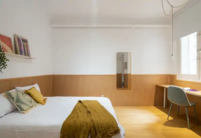 Camera da letto luminosa e ben arredata con letto matrimoniale in lenzuola bianche e dettagli color ocra; boiserie in legno, scrivania vicino alla finestra e specchio verticale completano lo spazio moderno e tranquillo.