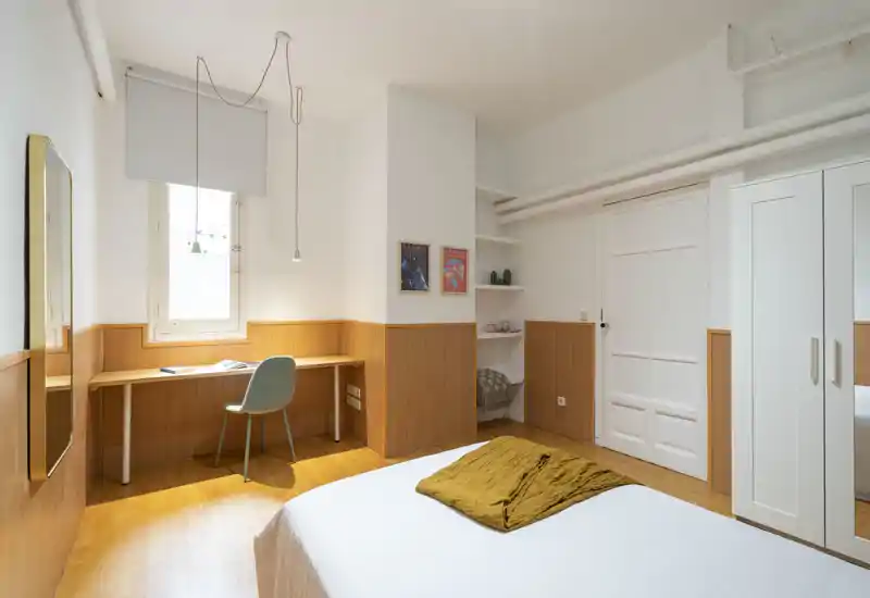 Bright, well-styled bedroom with a comfortable double bed, wooden paneling and a built-in desk by the window — ideal for rest and remote work.