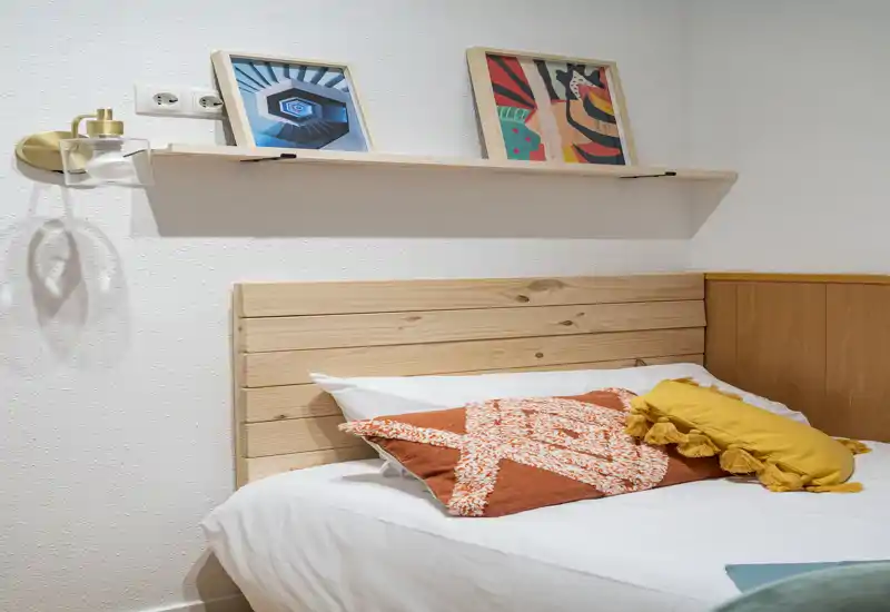Warm, well-styled bedroom corner with a wooden slat headboard, decorative pillows and framed artwork on a shelf — bright and inviting for short-term rentals.
