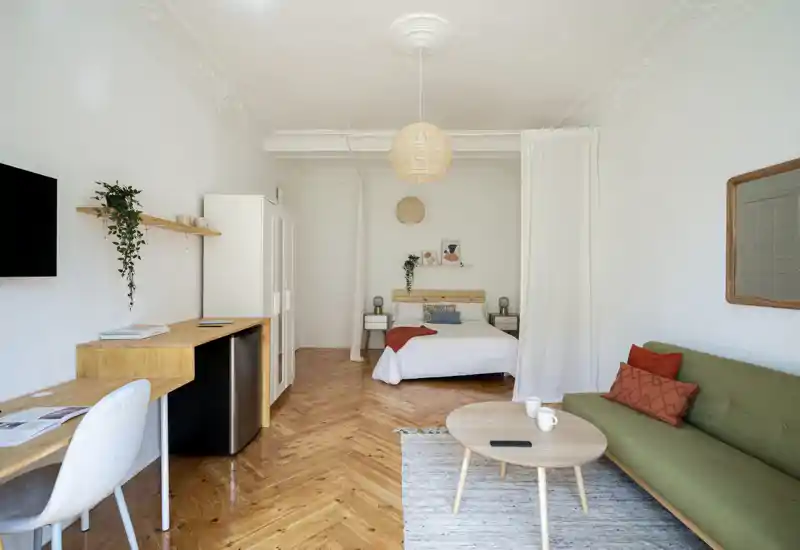 Bright, stylish studio bedroom with a central double bed, bedside tables and tasteful natural-wood furnishings. The space features a cozy seating area, workspace, and warm herringbone parquet flooring — ideal for an attractive short-term rental listing.