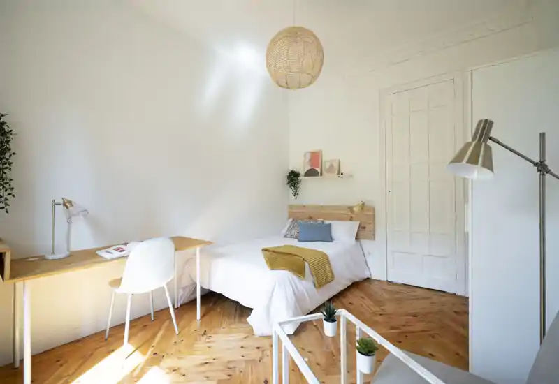 Bright, minimal bedroom with a comfortable double bed, wooden headboard and cozy textiles; a desk by the window and warm wooden floors complete the calm, well-lit space.