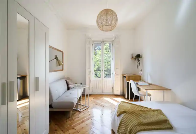 Bright, airy bedroom with wooden floors, a neatly made bed with a mustard throw, a small sofa, and a desk by the window — ideal for a single occupant or short stays.