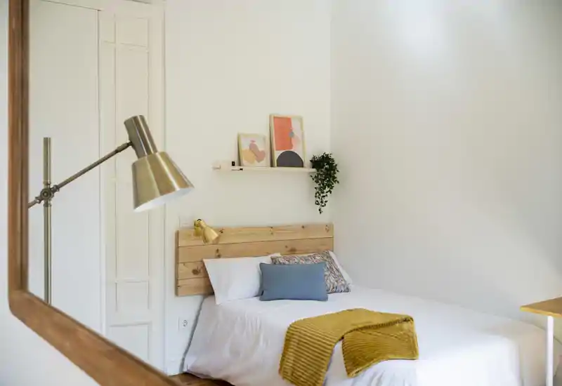 Bright, minimal bedroom with a wooden headboard, crisp white linens, decorative cushions and a mustard throw; pleasant natural light and tasteful wall decor.
