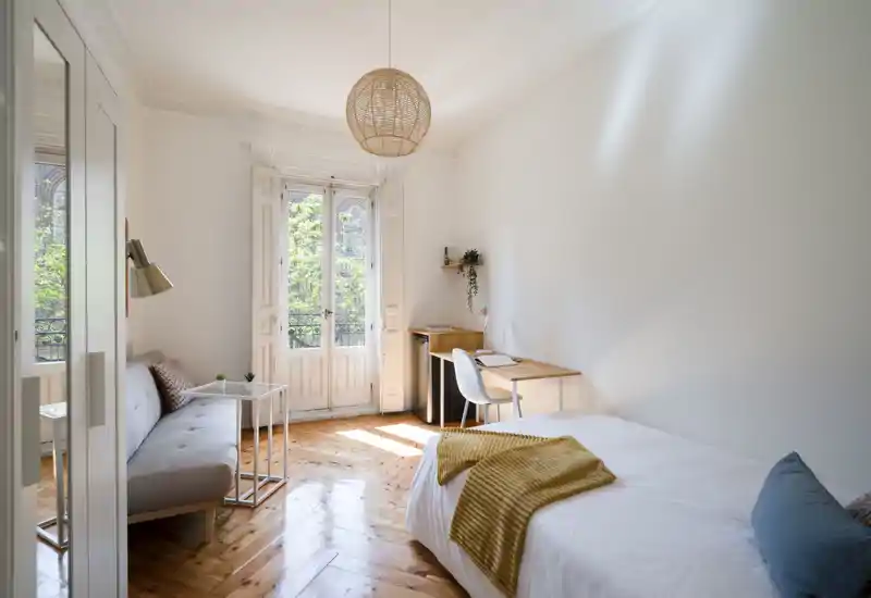 Bright, cozy bedroom with a comfortable single bed, warm wooden floors and a small workspace by a large window letting in abundant natural light.